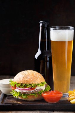 Burger, beer in a bottle and in a glass mug, french fries, sauce on a dark background, fast food concept.