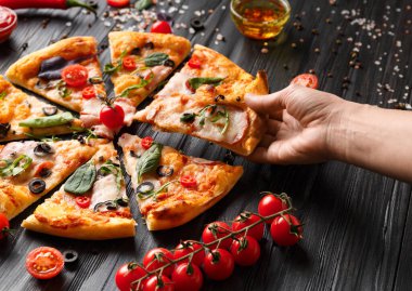 Eller, koyu ahşap arka planda kesilmiş pizzadan dilimler alır. Salamlı pizza, kurutulmuş et, sos, baharat, siyah arka planda taze vişneli domates. Kadınların elleri pizzayı paylaşır..