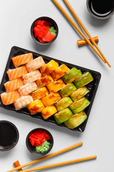 A set of bright multi-colored sushi rolls with shrimp, salmon, avocado in plastic packaging, Chinese chopsticks, sauce, ginger on a white background, top view. Sushi delivery