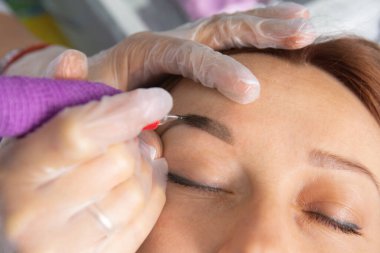 Beauty master performs permanent eyebrow makeup in a beauty salon, close-up. Hands of a cosmetologist doing microblading of eyebrows. Tattoo on the face. clipart