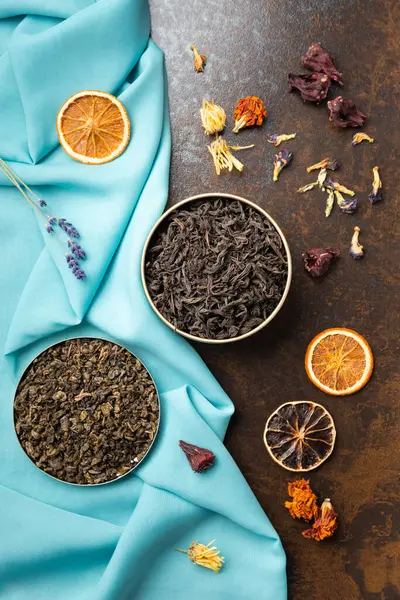 Composition of black, green, flower tea and dried orange slices on blue and brown backgrounds top view. Large-leaf tea, dried flowers and citrus chips on contrasting backgrounds.