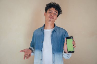 Genç kıvırcık adam kot giyip yeşil ekranlı akıllı telefonu tutuyor. Gülümse, şerefe ve mutlu bir ifade. Bu fotoğraf, erkek ifadesi reklamcılığı ve moda yaşam tarzı için uygun..