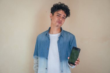 Genç kıvırcık adam kot giyip yeşil ekranlı akıllı telefonu tutuyor. Gülümse, şerefe ve mutlu bir ifade. Bu fotoğraf, erkek ifadesi reklamcılığı ve moda yaşam tarzı için uygun..