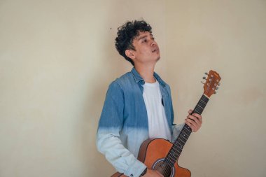 Genç kıvırcık adam gitar çalarken kot pantolon giyiyor. Gülümse, mutlu ol ve tezahürat yap. Bu fotoğraf, erkek ifadesi reklamcılığı ve moda yaşam tarzı için uygun..