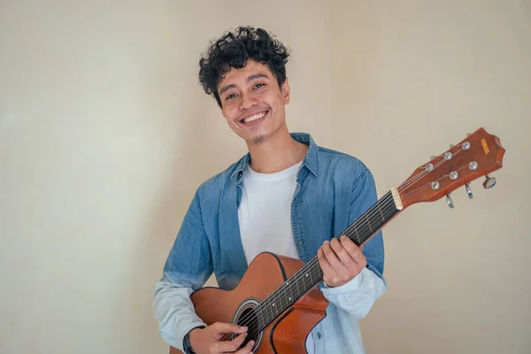 Young curly man wear denim clothes playing guitar. Smile, happy and cheers expression. The photo is suitable to use for man expression advertising and fashion life style.