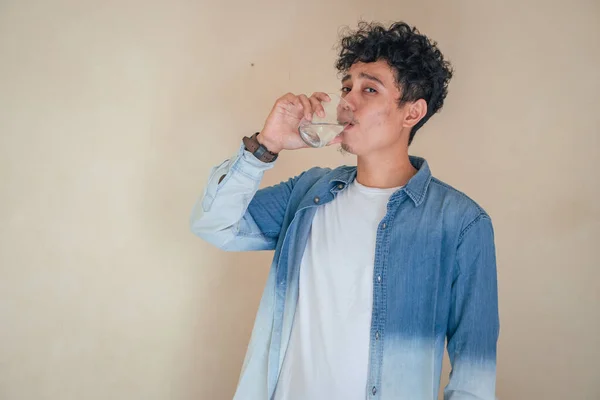 Young curly man with denim clothes hold a glass water. Smile, cheers and happy expression. The photo is suitable to use for mineral water bottle promotion and healthy life advertising.