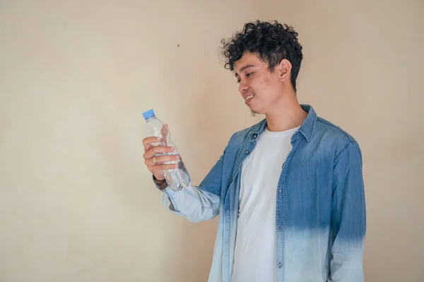 Young curly man with denim clothes hold a bottle water. Smile, cheers and happy expression. The photo is suitable to use for mineral water bottle promotion and healthy life advertising.