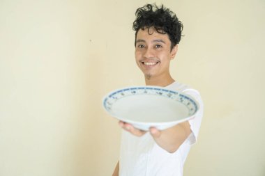 Genç kıvırcık adam beyaz tişörtler giyer. Tabak, kaşık ve çatal tutar ve jest yapar. Fotoğraf sağlıklı bakım ve günlük etkinlik içeriği medyası için kullanılabilir.