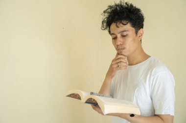 Kıvırcık genç adam kitap okur ve tutar beyaz kıyafetler giyer. Fotoğraf eğitim içeriği medya ve ifade reklamları için kullanılabilir.