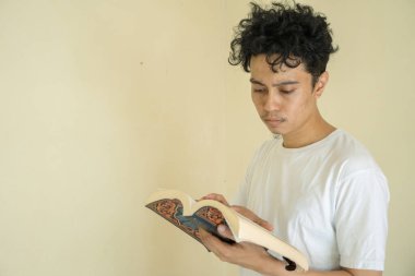 Kıvırcık genç adam kitap okur ve tutar beyaz kıyafetler giyer. Fotoğraf eğitim içeriği medya ve ifade reklamları için kullanılabilir.