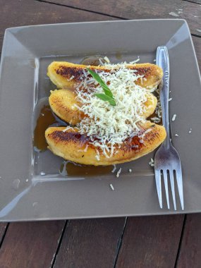 Gri tabakta ve çatalda karamelli peynirli ızgara muz. Fotoğraf yemek arkaplanı, poster ve yiyecek içeriği medyası için kullanılabilir.