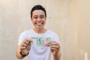 Endonezyalı erkekler mutlu olur ve parayı tutarken beyaz tişört giyer. Bu fotoğraf, erkek ifadesi reklamcılığı ve moda yaşam tarzı için uygun..