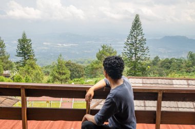 Adam çitin yanında oturuyor ve manzara dağına bakıyor. Fotoğraf sakin hareketler, boş zaman aktiviteleri ve park geçmişi için kullanılabilir.