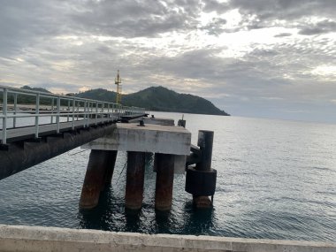 Çelik destekli beton bir iskele, sakin bir denize uzanır. Arka planda ufuk ve gökyüzü gri bulutlarla dolu bir sıradağdır.