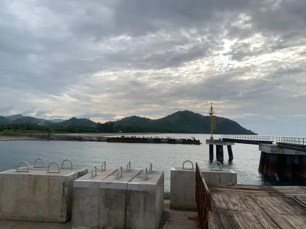 Bulutlu bir gökyüzü, ahşap bir iskele, bir tekne iskelesi ve arka planda dağlar olan bir liman manzarası..