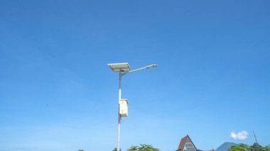 Açık mavi gökyüzüne karşı dik duran güneş enerjili bir sokak lambası, açık havada aydınlatma için yenilenebilir enerji kullanımını vurguluyor.