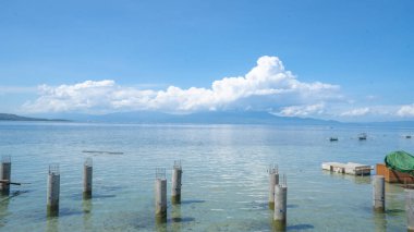 Uzak dağlar ve sakin mavi gökyüzü manzaralı pitoresk bir deniz manzarası. Görüntüde sakin sular ve berrak bir ufuk var, sakin bir atmosfer yaratıyor. Bazı beton sütunlar kısmen suyun altında duruyor. Bu da bir yoruma işaret ediyor.