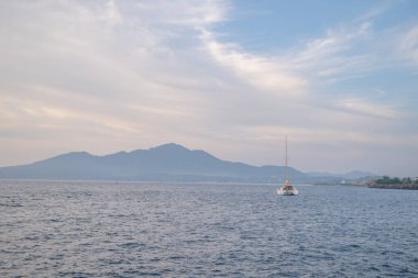 Yalnız bir yelkenli, sakin bir okyanusta, dağlık bir kıyı şeridine doğru yol alır..