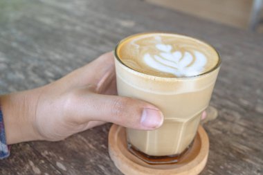 Camdan bir fincanda latte ile lattenin yakın plan görüntüsü. Kahve bir el tarafından tutuluyor, ahşap bir masada dinleniyor..