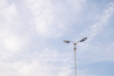 Tek bir sokak lambası beyaz bulutlarla dağılmış açık mavi bir gökyüzünün arka planına karşı dimdik ayakta duruyor. Işık direği dizaynda basittir, aşağı bakan iki lamba aşağıdaki sokağı aydınlatmaya hazırdır..