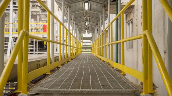 Sarı bir tırabzan geçidi endüstriyel bir alana çıkıyor. Geçit metalden yapılmış ve ızgara deseni var..