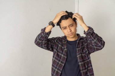 Ekose gömlekli bir adam yüzünde endişeli bir ifadeyle saçına dokunuyor. Beyaz bir duvarın karşısında duruyor, aşağıya bakıyor ve düşüncelere dalmış..