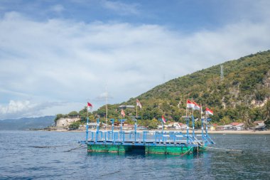 Bu görüntüde renkli bir feribot iskelesi var. Resimli bir sahil köyünde yer alıyor. Endonezya bayraklarıyla süslenmiş rıhtım, yemyeşil tepeler ve dağlardan oluşan yemyeşil bir zemin ile turkuaz sulara kadar uzanıyor..