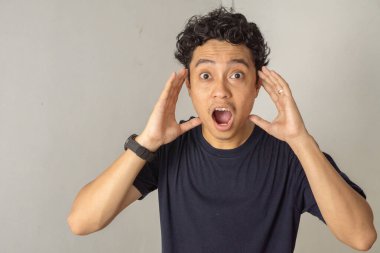 Ağzı açık, şaşırmış bir gencin portresi. Elleri kulaklarına yakın bir şekilde kameraya bakıyor, bir şok ya da şaşkınlık jesti gösteriyor..