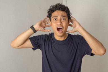 Şok olmuş bir yüz ifadesi olan bir adam elleriyle kulaklarını kapatır. Görüntü, yoğun bir şaşkınlık, korku ya da inançsızlık anını yakalar. Sıradan bir tişört giymiş..