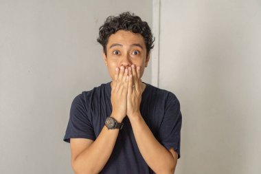 Kıvırcık saçlı genç bir adam kameraya şaşırmış bir ifadeyle bakıyor, elleriyle ağzını kapatıyor. O, mavi bir tişört ve bir saat giyiyor. Arka plan beyaz bir duvar..