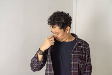 Genç bir adam beyaz bir duvara karşı duruyor, burnunu eliyle tutuyor ve aşağıya bakıyor. Ekose bir gömleğin altına siyah bir tişört ve bileğinde bir saat giyer..