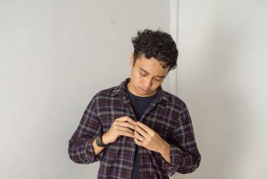 Ekose gömlekli genç bir adam beyaz bir stüdyoda dikiliyor, düğmelerini ilikliyor, aşağıya bakıyor..