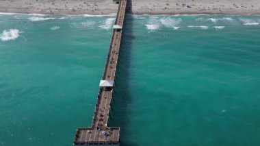 This 4K aerial drone footage offers a stunning view of a Florida pier, coastal beach, and ocean. The footage captures the natural beauty of the area, showcasing the crystal-clear water and the picturesque pier jutting out into the ocean.