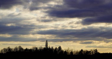 Güneşin bulutlu bir gökyüzü ve karanlık bir manzara siluetiyle battığı Kuzey Ren Vestfalya manzarası.
