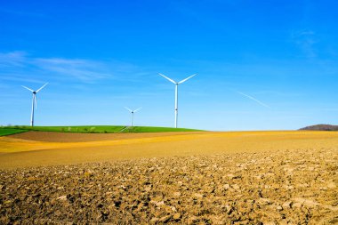 Mavi gökyüzüne karşı yel değirmeni ve önünde bir tarla. Rüzgar enerjisiyle enerji üretimi.