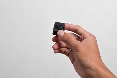 Elinde beyaz arka planda izole edilmiş bir casus kamerası var. Siyah güvenlik kamerası, Mini casus kamerası.