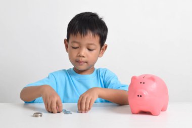 Asyalı küçük çocuk, beyaz arka planda izole edilmiş pembe kumbaralarda para biriktiriyor. Çocuk konsepti için para biriktiriyorum. Tasarruf etmeyi öğren. Para, finans ve sigorta kavramı