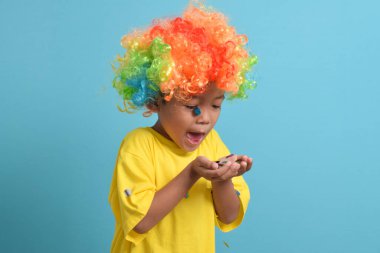 Palyaço peruğu takmış şirin Asyalı çocuk mavi arka planda izole edilmiş parti konfetileri üflüyor. Boşluğu kopyala