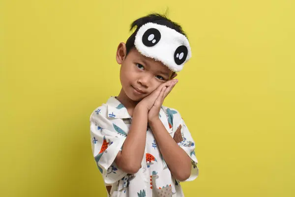 stock image Portrait of cute Asian child wearing pajamas and sleep mask putting his hand under his cheek isolated on yellow background. Child wants to sleep. Healthy sleep concept