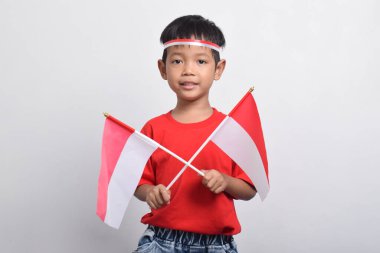 Kırmızı tişörtlü mutlu bir Asyalı çocuk elinde iki bayrak ve gülümsüyor, beyaz arka planda soyutlanmış. Endonezya Bağımsızlık Günü konsepti.
