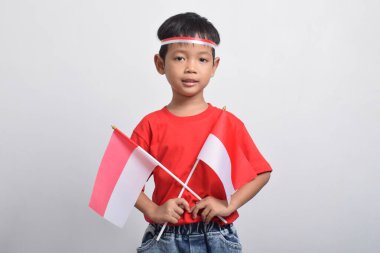 Kırmızı tişörtlü mutlu bir Asyalı çocuk elinde iki bayrak ve gülümsüyor, beyaz arka planda soyutlanmış. Endonezya Bağımsızlık Günü konsepti.