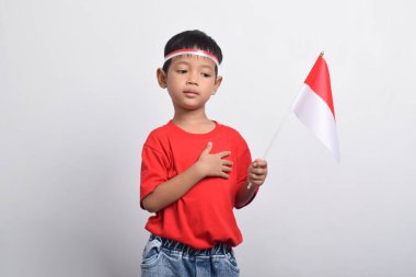 Kırmızı tişörtlü mutlu bir Asyalı çocuk, beyaz arka planda izole edilmiş, eli göğsünde bir bayrak tutuyor. Endonezya Bağımsızlık Günü konsepti.