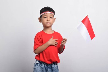 Kırmızı tişörtlü mutlu bir Asyalı çocuk, beyaz arka planda izole edilmiş, eli göğsünde bir bayrak tutuyor. Endonezya Bağımsızlık Günü konsepti.