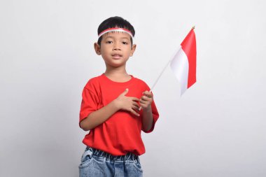 Kırmızı tişörtlü mutlu bir Asyalı çocuk, beyaz arka planda izole edilmiş, eli göğsünde bir bayrak tutuyor. Endonezya Bağımsızlık Günü konsepti.