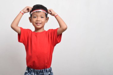 Kırmızı tişört giyen mutlu Asyalı çocuk ve beyaz arka planda izole bir şekilde kafasını tutan kırmızı ve beyaz saç bandı. Endonezya Bağımsızlık Günü konsepti.