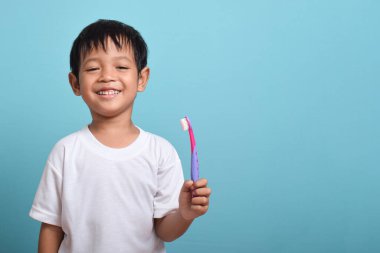 Mavi arka planda diş fırçası tutan mutlu bir Asyalı çocuk. Çocuk dişlerini göstererek gülümsüyor. Çocuk diş bakımı ve hijyen