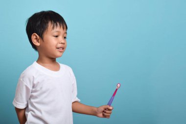 Mavi arka planda diş fırçası tutan mutlu bir Asyalı çocuk. Çocuk dişlerini göstererek gülümsüyor. Çocuk diş bakımı ve hijyen