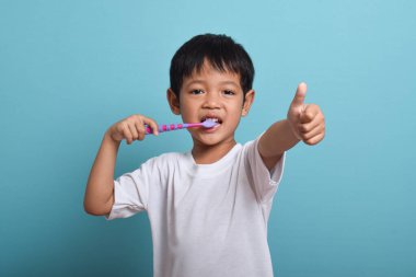 Kameraya başparmağını uzatırken dişlerini fırçalayan mutlu bir Asyalı çocuk. Mavi arka planda izole edilmiş. Sağlık, ağız temizliği.