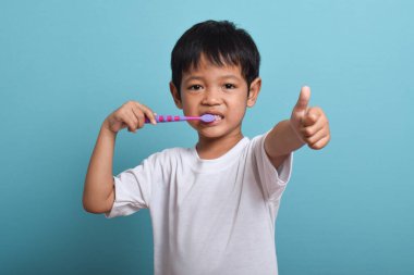 Kameraya başparmağını uzatırken dişlerini fırçalayan mutlu bir Asyalı çocuk. Mavi arka planda izole edilmiş. Sağlık, ağız temizliği.