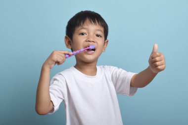 Kameraya başparmağını uzatırken dişlerini fırçalayan mutlu bir Asyalı çocuk. Mavi arka planda izole edilmiş. Sağlık, ağız temizliği.
